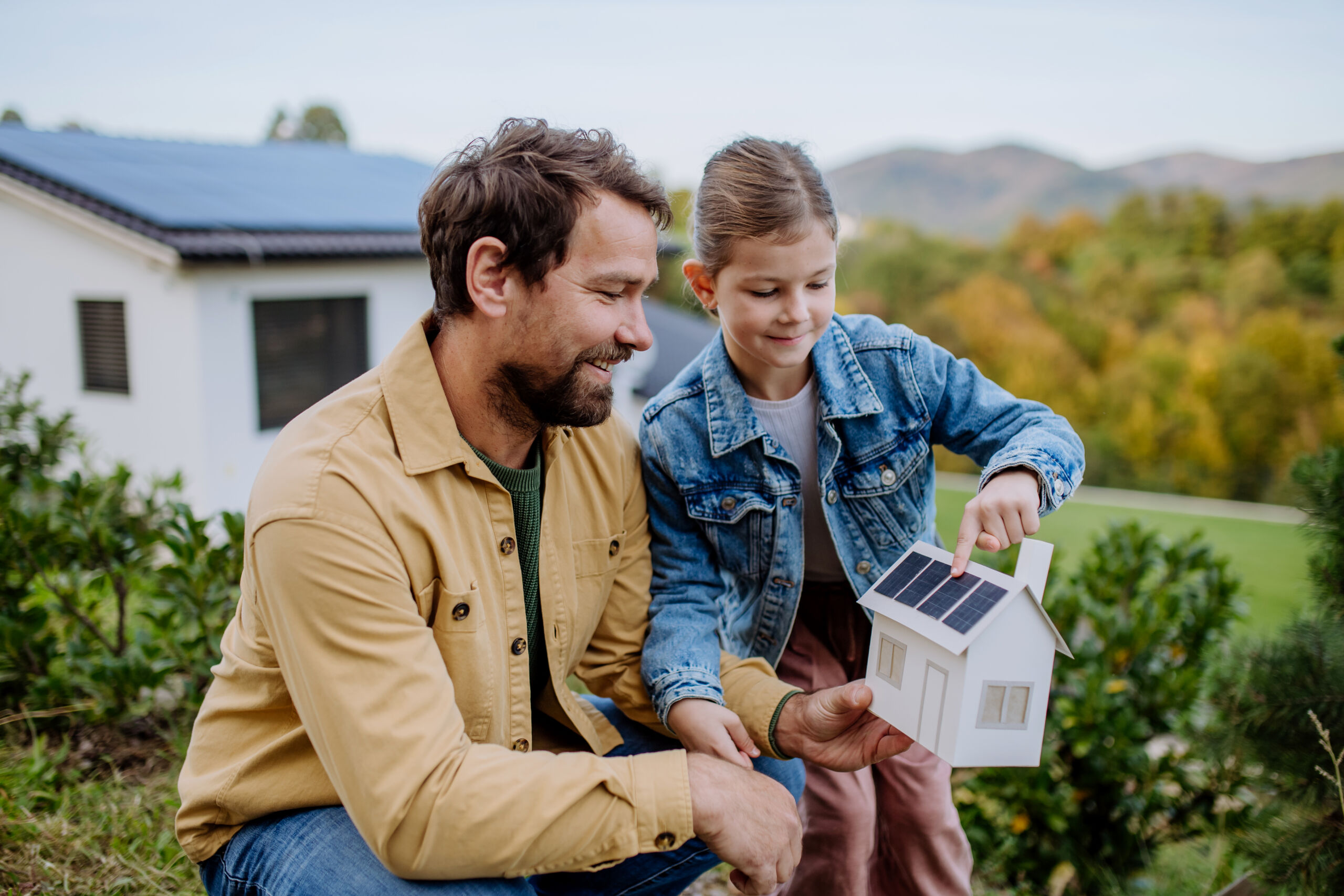 An image of a father and his daughter. She is showing him a small model house with solar panels on the roof.