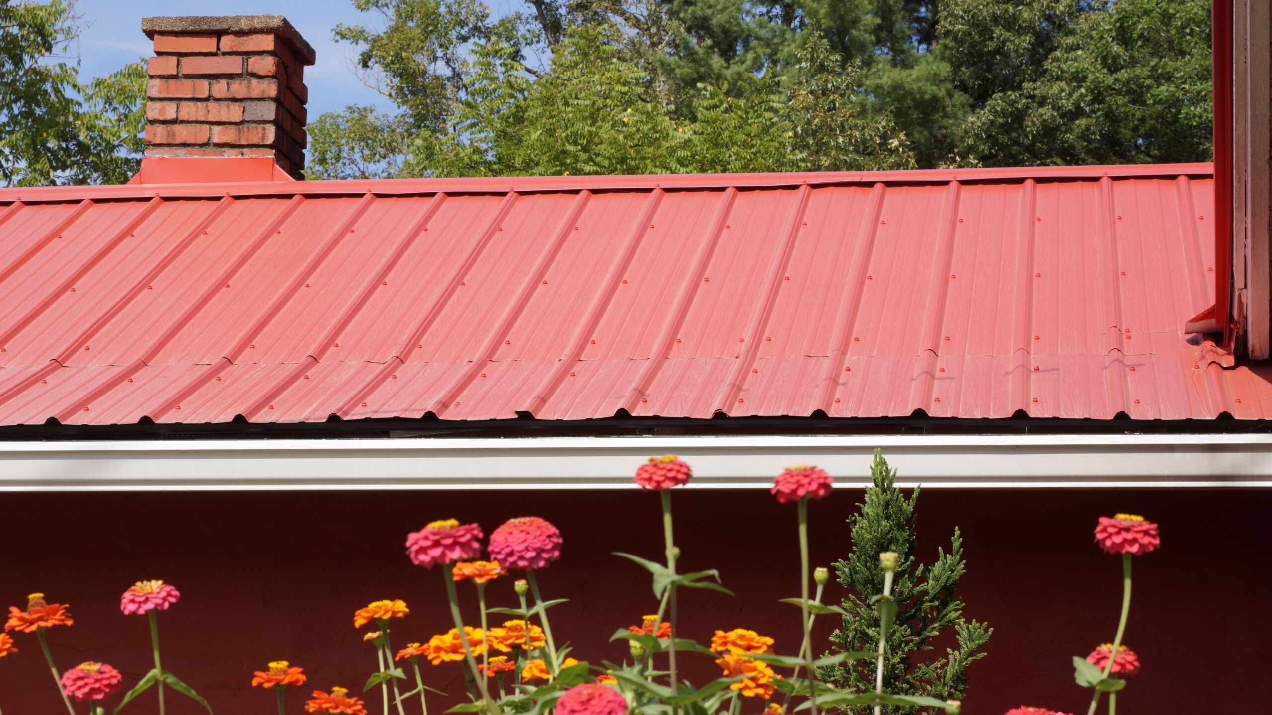 An image of a beautiful red roof on a house with a flower garden in the foreground. Do Metal Roof Save Money?