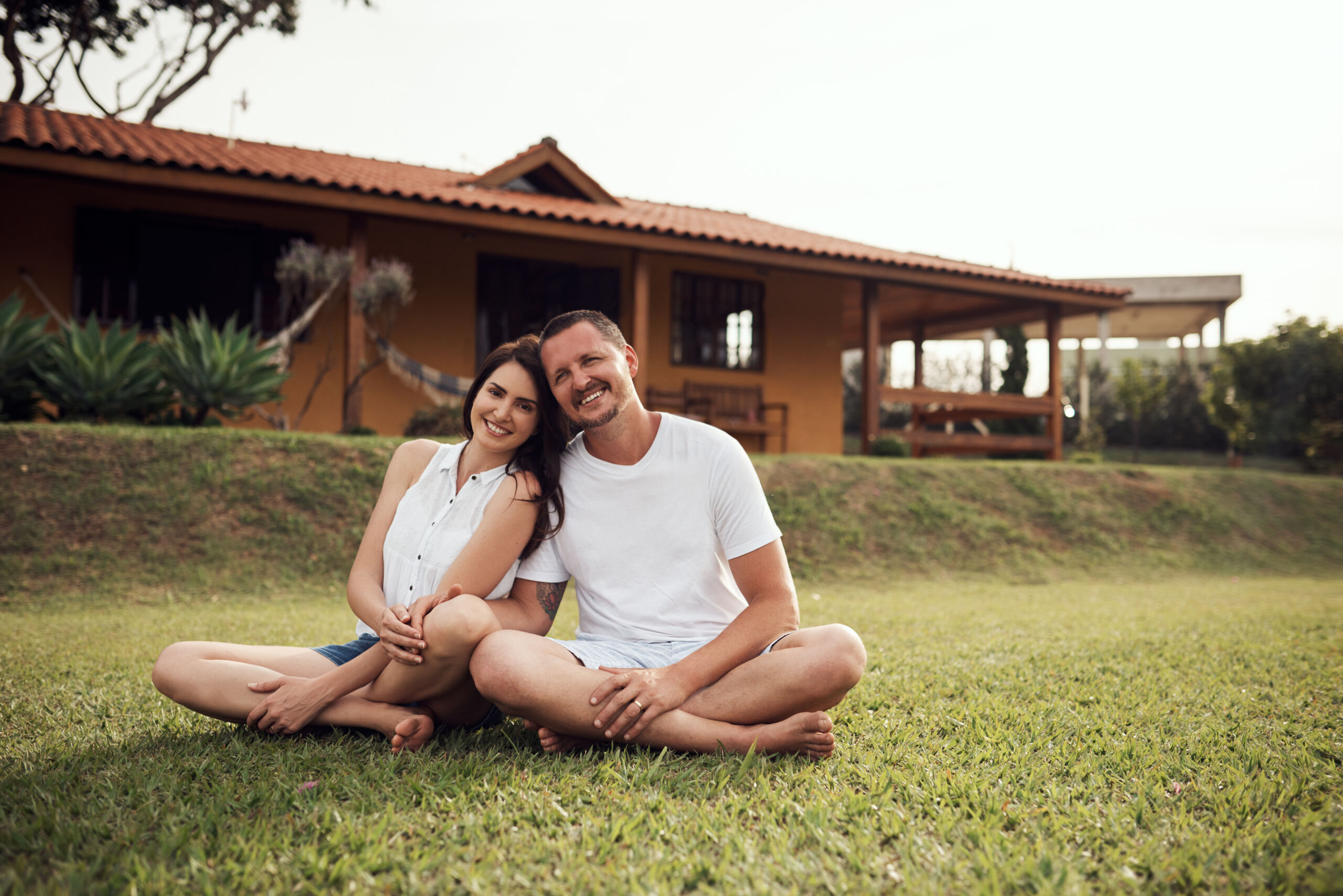 An image of a young, married couple sitting in the grass in front of their house. Both are smiling. Did insurance pay for replacing their roof?
